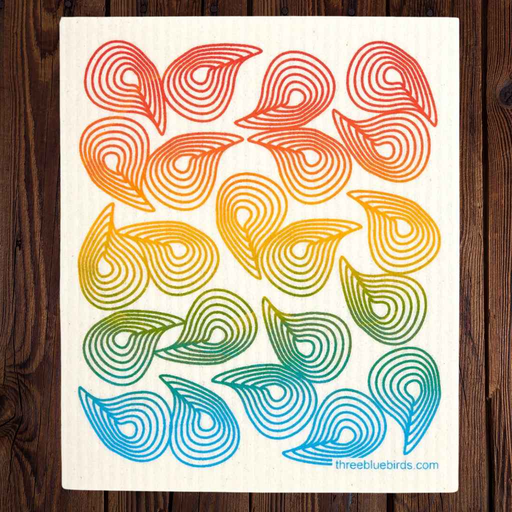 A square swedish dish towel featuring a retro pattern of concentric wavy lines in a rainbow gradient, laid flat on a dark wood background. The print has a white background and the website threebluebirds.com