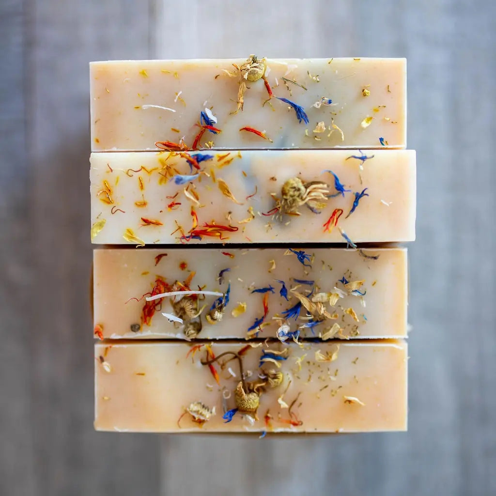 Four rectangular bars of handmade soap stacked vertically, featuring a pale peach-beige base with dried botanical inclusions in orange, blue, and gold tones, set against a neutral gray background.