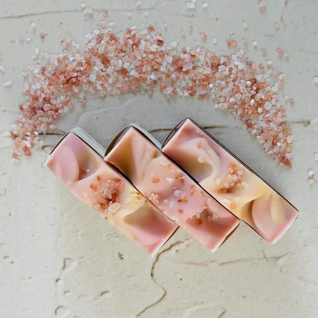 Three rectangular pink and cream soap bars with embedded coarse salt crystals, arranged on a light beige textured surface with a scattered halo of pink salt above them.