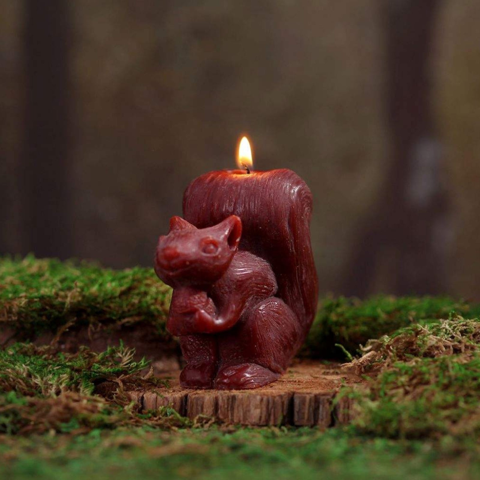 A lit squirrel-shaped candle in a dark reddish-brown color, placed on a small piece of wood amidst green moss in an outdoor forest-like setting.