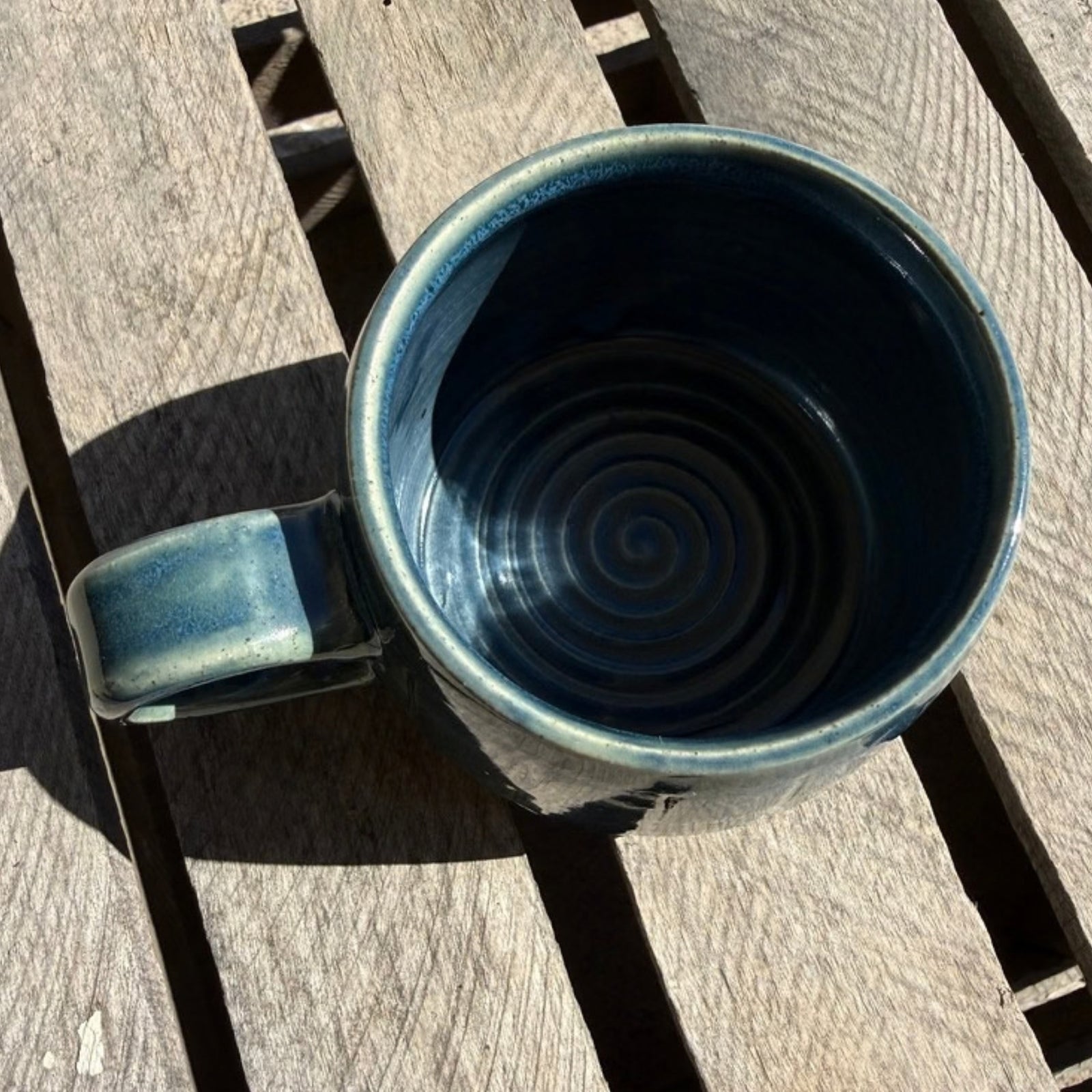 A handmade ceramic mug with a dark blue spiral-glazed interior and a matching handle, shown from a top-down perspective on weathered wooden deck slats.