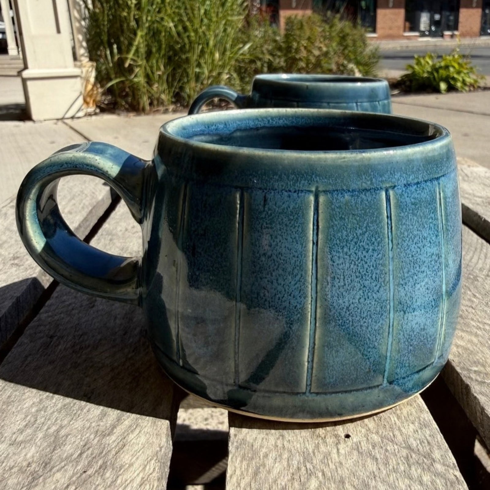 A large ceramic mug with a textured blue glaze and vertical fluted detailing, sitting on a wooden outdoor table with another mug in the background.