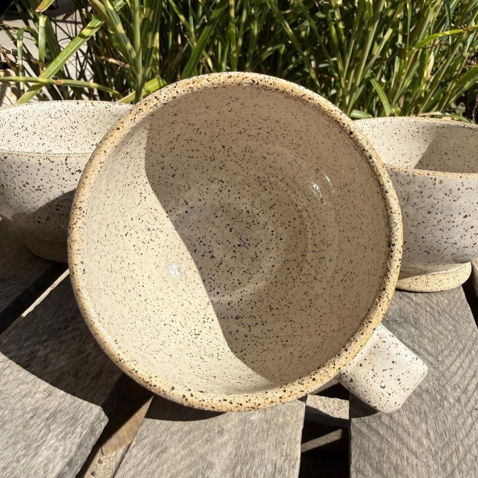 A speckled ceramic mug with a handle, shown in warm natural sunlight on a weathered wooden deck with green plants in the background.