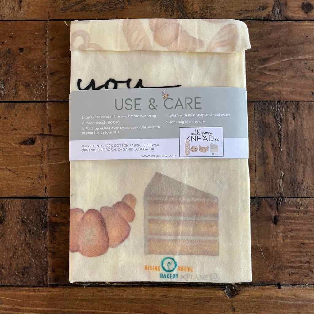 Back side of Beeswax bread bag with paper label showing care instructions, illustrations of bread and cookies on bag, all on a rustic wooden surface