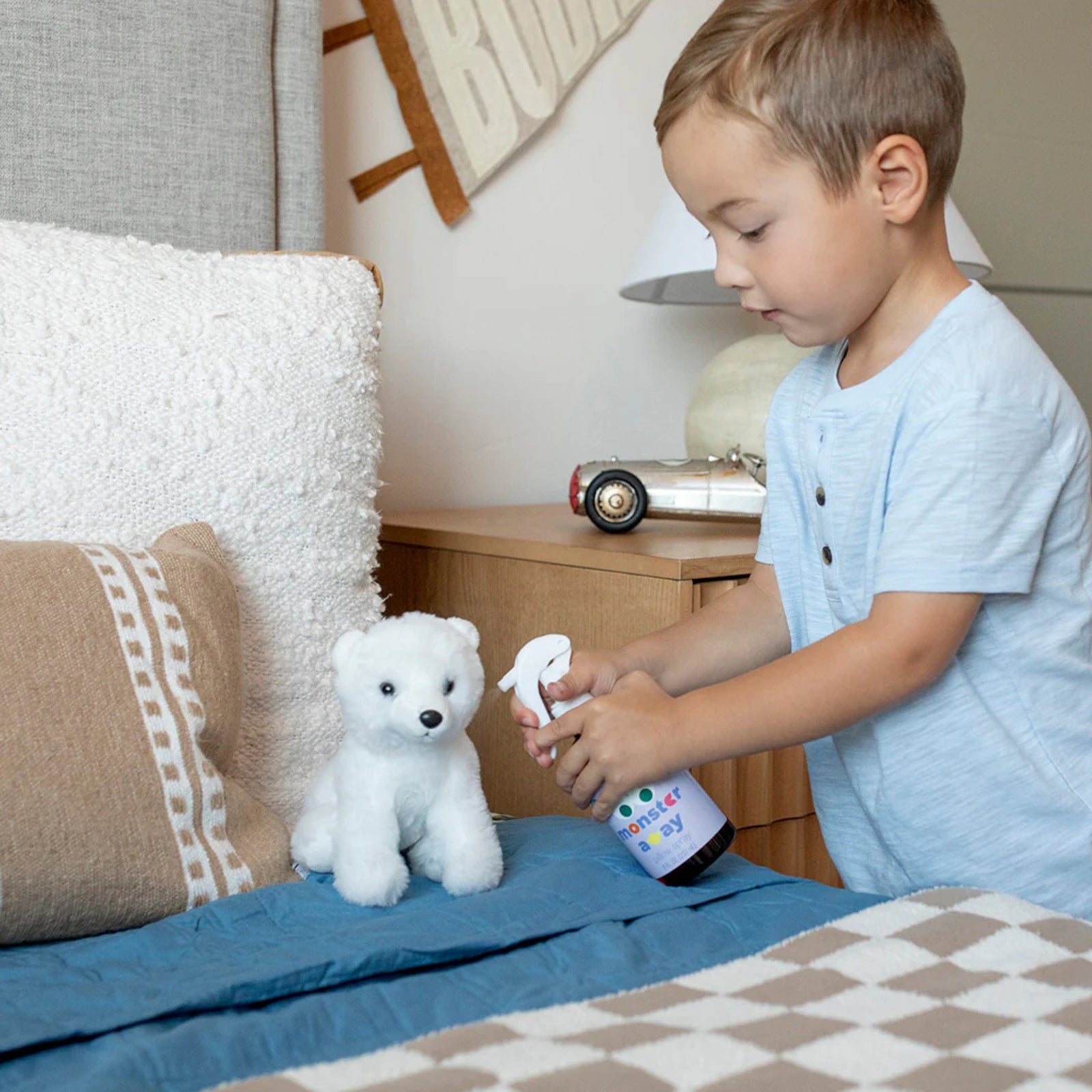 A young boy in a light blue shirt sprays a white polar bear plush toy with a small spray bottle in a child's bedroom setting.