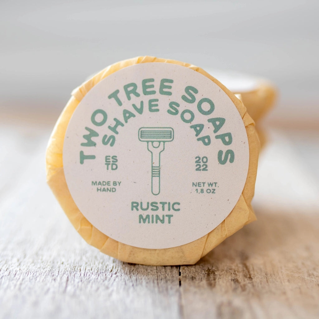 A wrapped bar of Two Tree Soaps Shave Soap in Rustic Mint, resting on a light wooden surface with a soft-focus background.