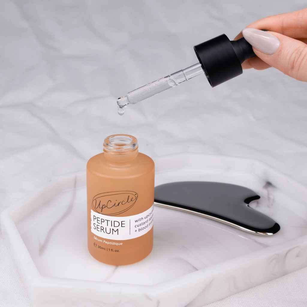 A hand with pale pink manicured nails holds a glass dropper above an open bottle of UpCircle Peptide Serum. The serum inside the clear bottle is a warm peachy nude shade, and a stainless steel gua sha massage tool rests on a white marble tray in the background.