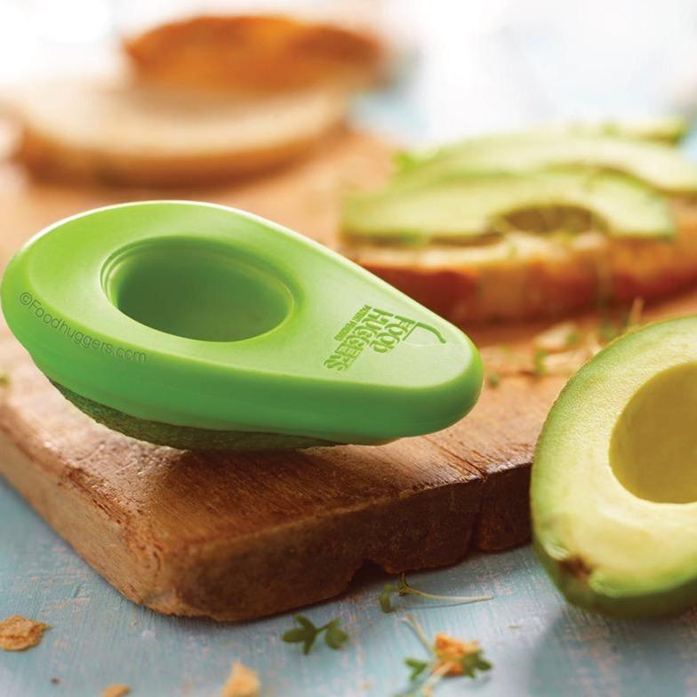 avocado hugger on ripe avocado, next to sandwich being make on wooden cutting board