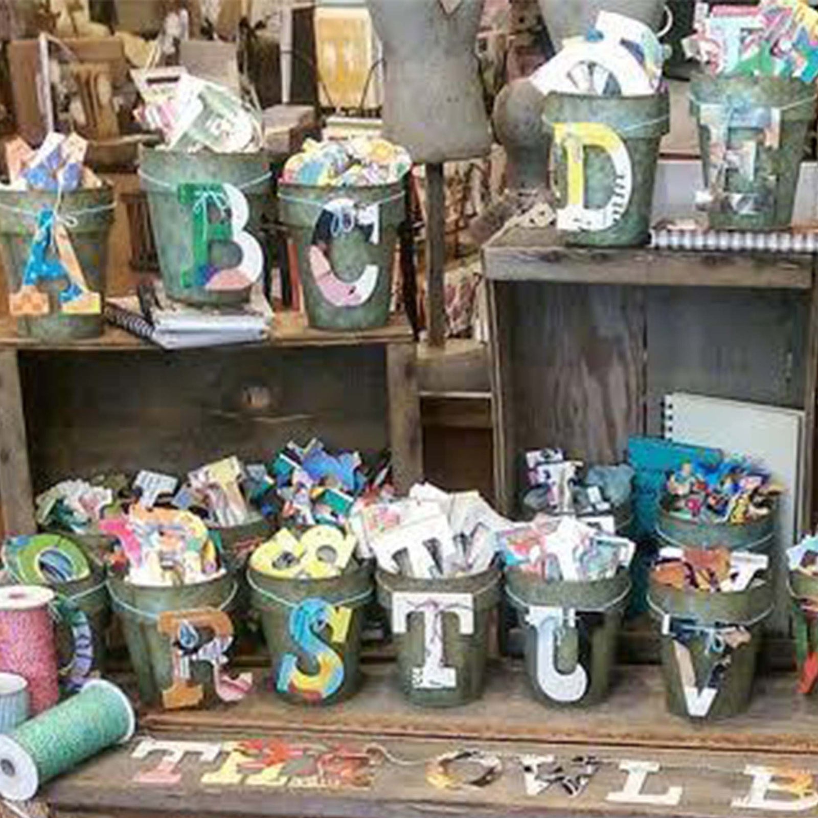 metal buckets placed on wooden crates  containing letters A-D and R-V, alongside spools of multicolored ribbon,  Each bucket is overflowing with a variety of designs of each letter.