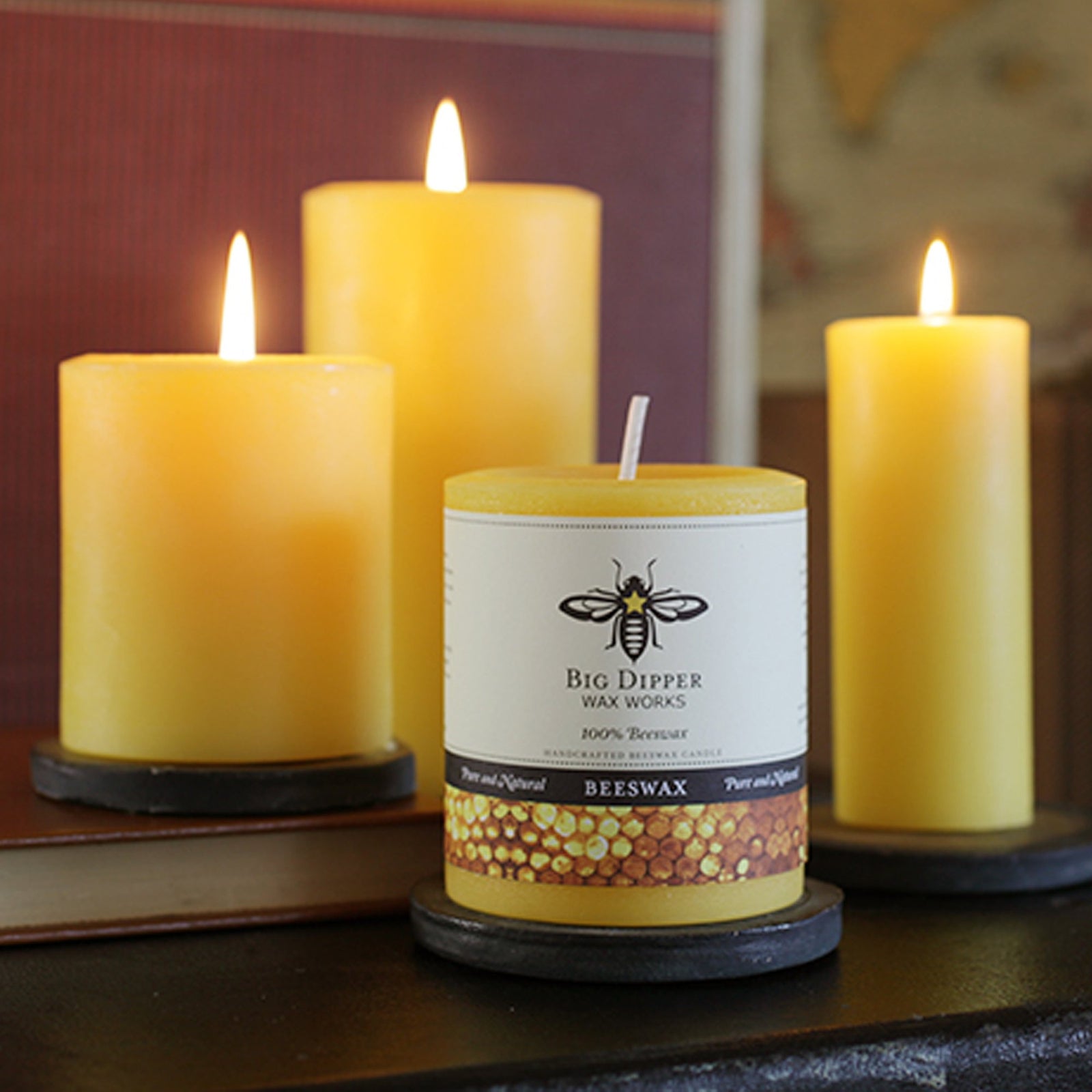 four 100% Pure Beeswax Candles of different heights and widths, lit atop on ebony table top including red leather bound books