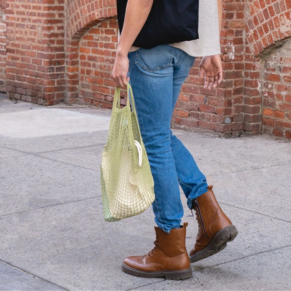 A person walking outdoors on a sidewalk in front of a red brick building, wearing blue jeans, brown ankle boots, a black top, and holding a light green mesh tote bag.