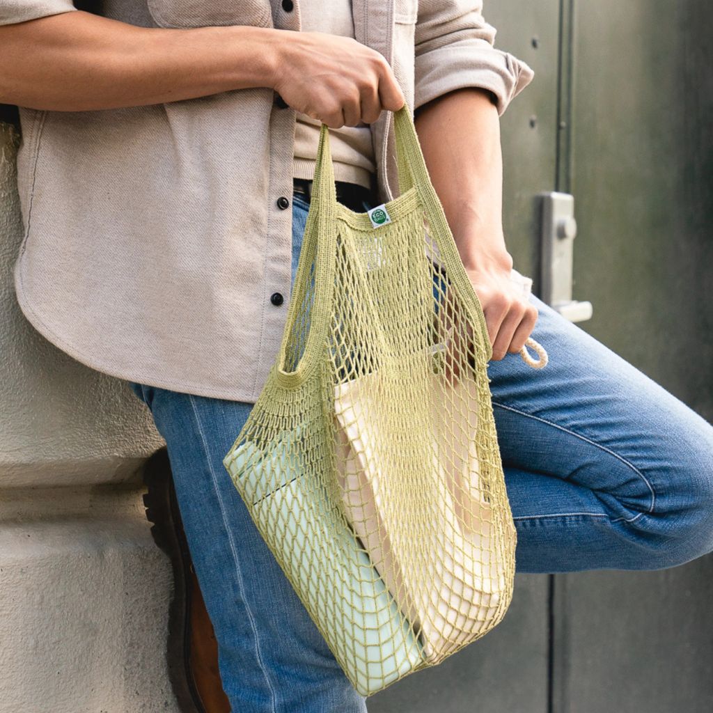 A person wearing a beige shirt and blue jeans holds a light green mesh reusable tote bag filled with items, standing against a dark metal door.
