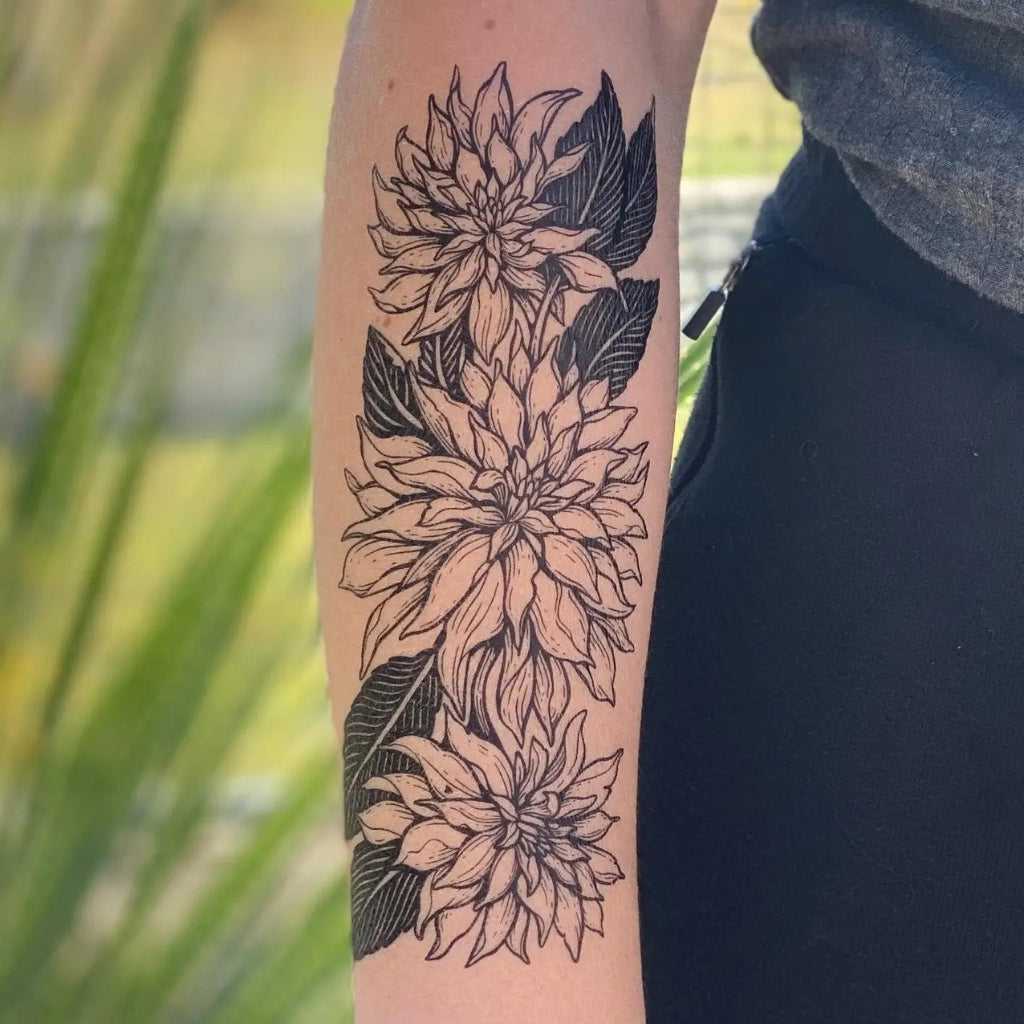 A close-up of a person's forearm featuring a black and white line art tattoo of three dahlia flowers and leaves, shown outdoors with a blurred green background.