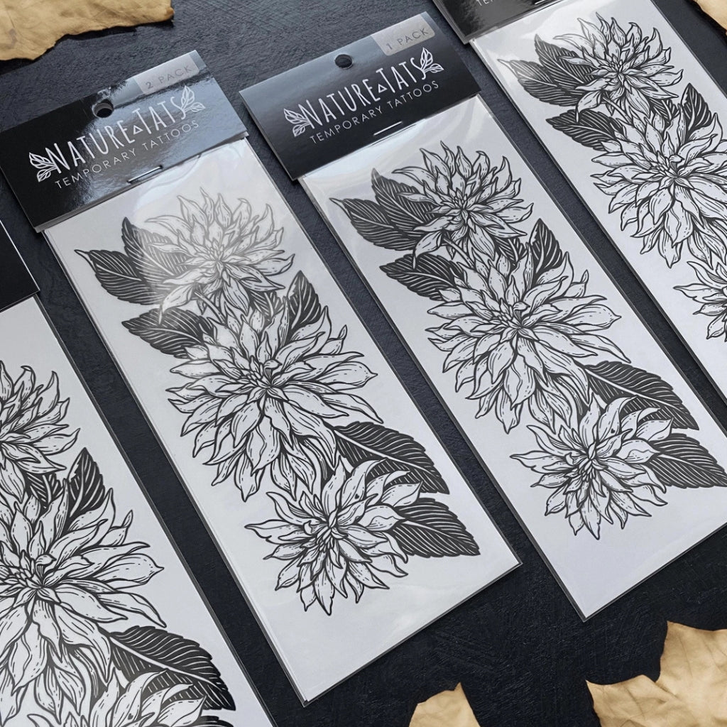 Three packages of Nature's Tattoos temporary tattoos featuring black and white dahlia flower designs, arranged on a dark textured surface with dried leaves.