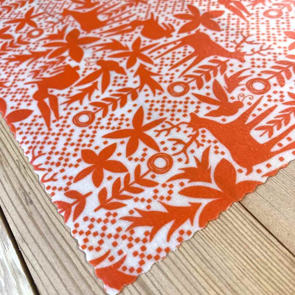 A close-up of a white beeswax fabric with an orange folk-art style print featuring deer, leaves, and geometric patterns, laid diagonally on a light wood surface.