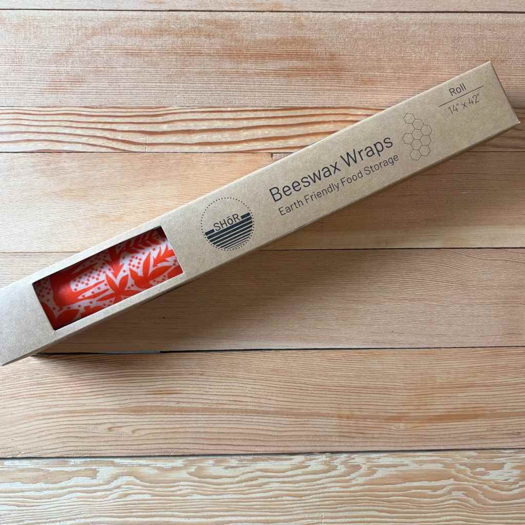 A roll of beeswax wraps in a kraft paper box with a clear window showing the red patterned wrap inside, placed on a light wood plank surface.