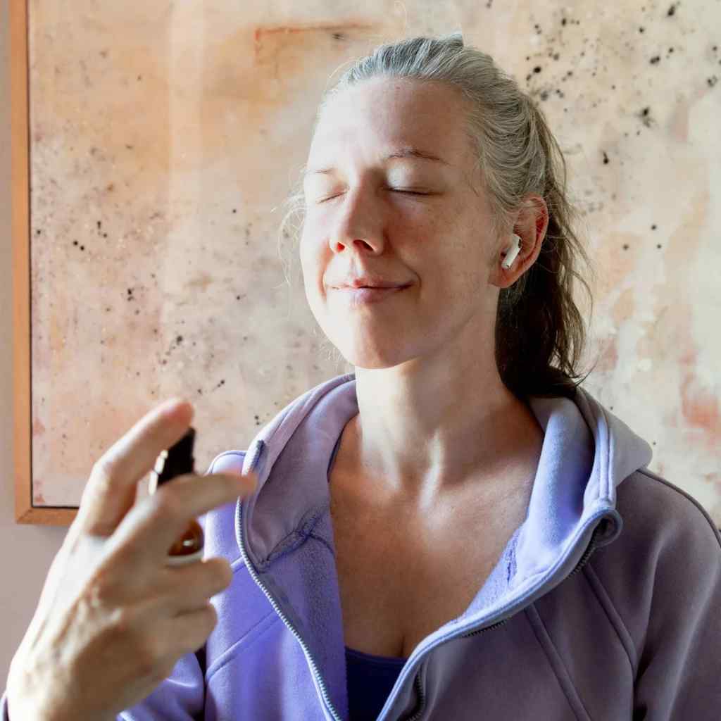 Woman in a purple hoodie spraying Mona Moon Natural's Chill Out Wellness Spray on herself in front of a neutral wall.