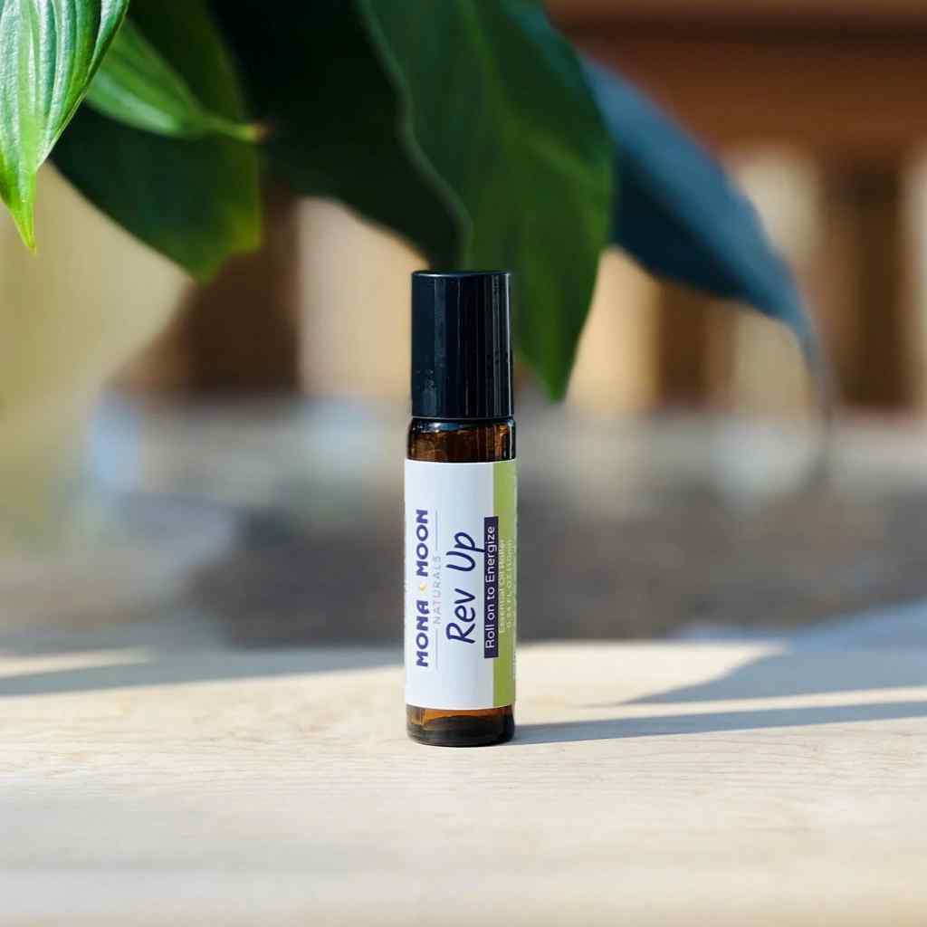 Roll-on bottle labeled 'Rev Up' by Mona Moon on a wooden surface with green leaves in the background.