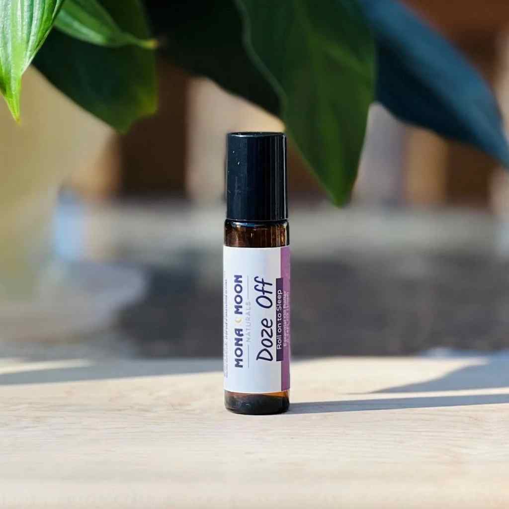 Roll-on bottle labeled 'Doze Off' by Mona Moon on a wooden surface with a blurred plant in the background.