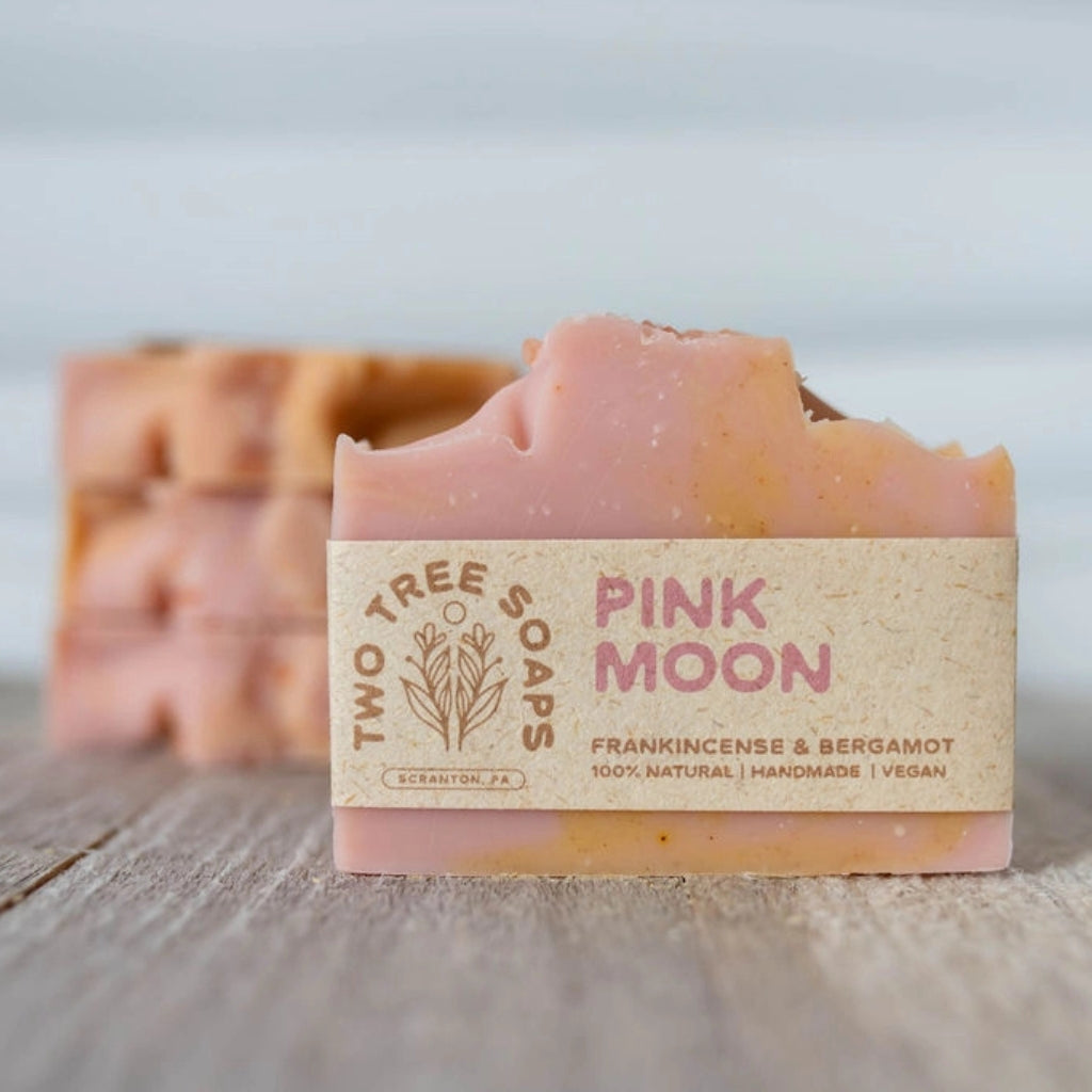 A bar of Two Tree Soaps 'Pink Moon' handmade soap in pink and beige, resting on a rustic wooden surface with a stack of similar soaps blurred in the background.