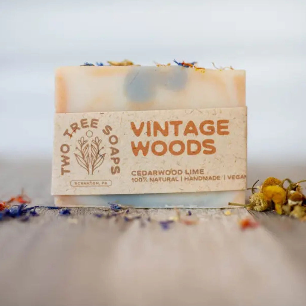 A rectangular bar of Two Tree Soaps 'Vintage Woods' soap in a natural beige paper wrapper, with dried flower petals on top, set on a rustic wooden surface with blurred dried flowers.
