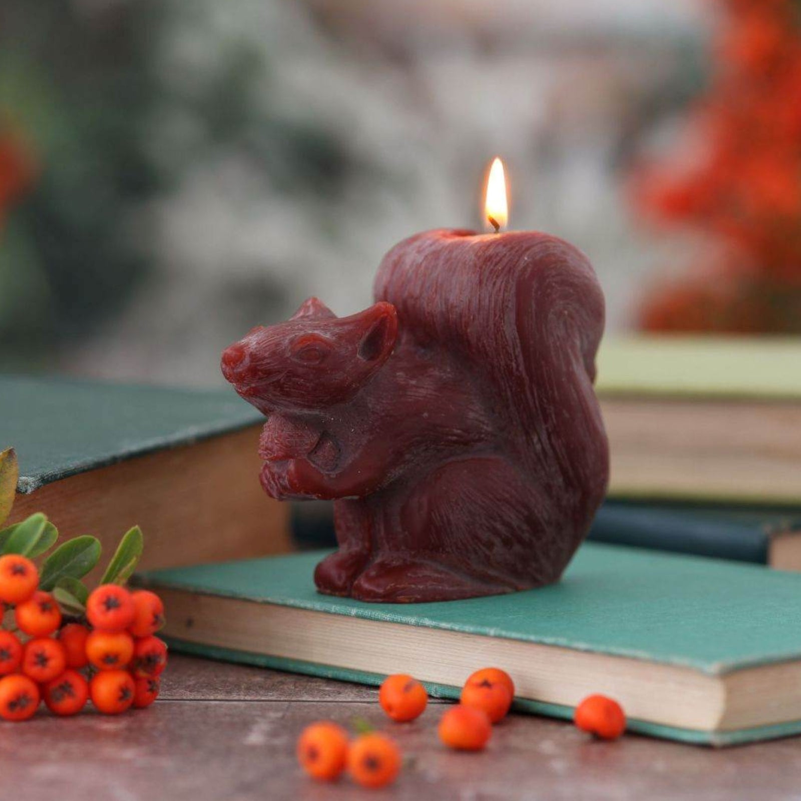 A sculpted squirrel-shaped candle in a dark reddish-brown wax sits lit on top of a green book, surrounded by autumn-themed props including small orange berries and stacked books in a softly blurred outdoor setting.