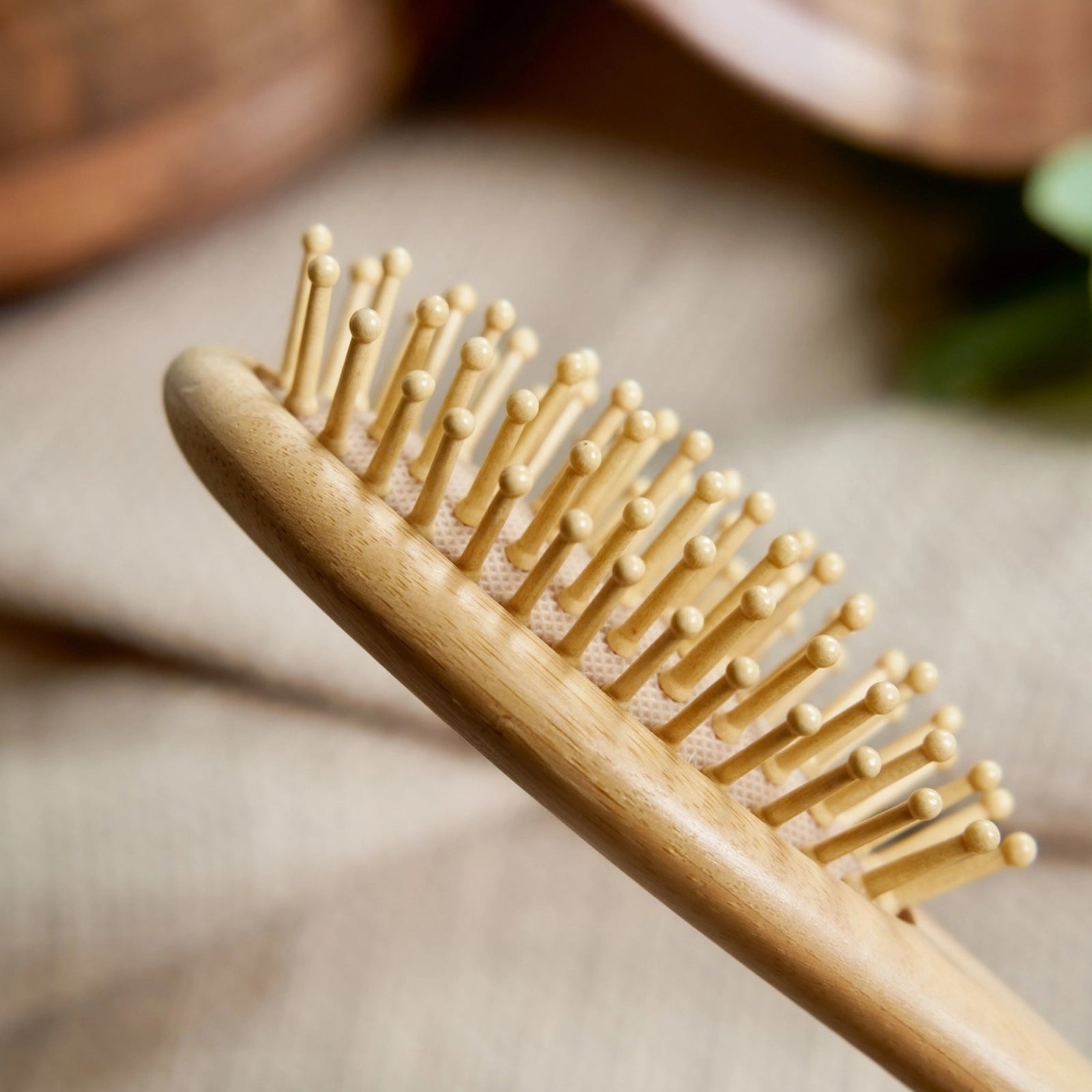 A close-up of a light natural wood hairbrush with soft bamboo bristles, resting on a textured beige fabric background.