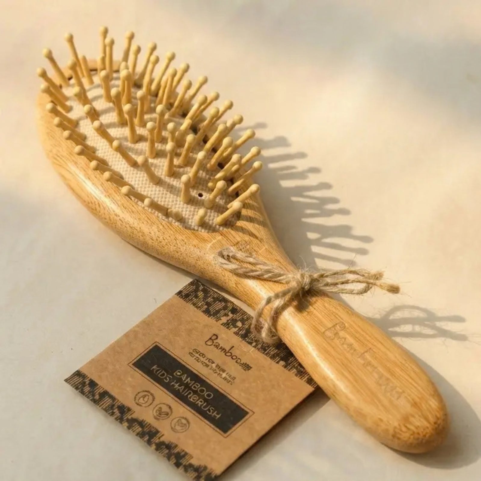 A natural bamboo hairbrush with light wood bristles and a matching wooden handle, tied with twine and shown with a kraft paper tag on a warm beige background.