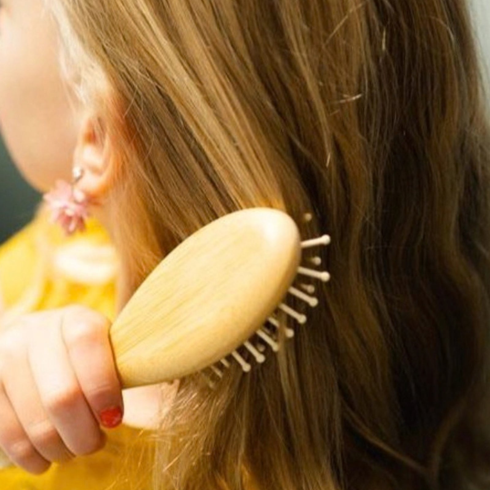 A close-up lifestyle shot of a person with long light brown hair using a light natural wood hairbrush to brush their hair.