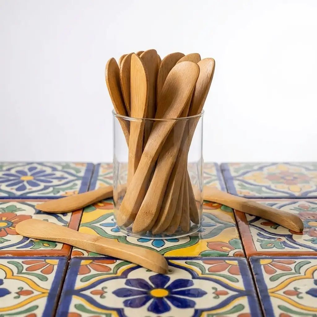 A set of light natural wood serving spoons in a clear glass jar, with a few spoons placed on a colorful patterned tile surface against a white background.