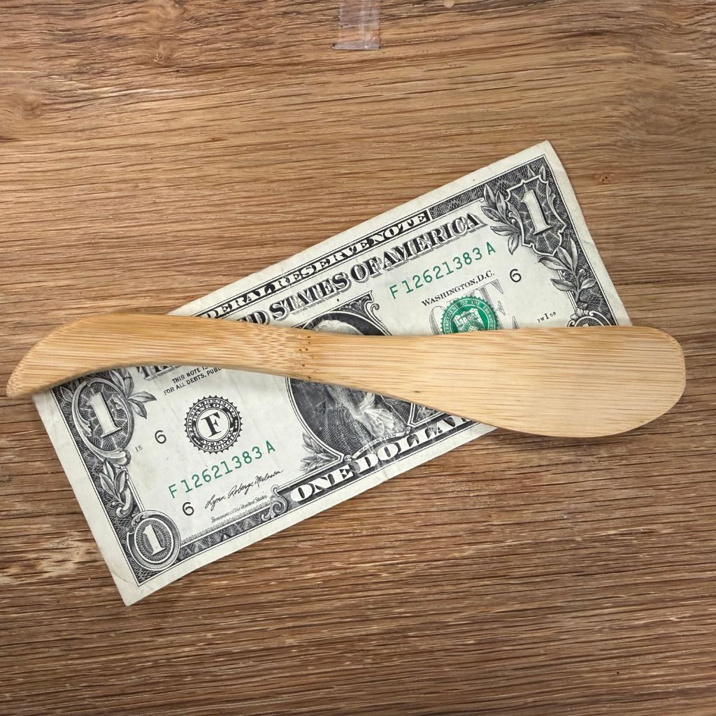 A light natural wood spreader tool rests diagonally across a U.S. one-dollar bill on a wooden tabletop.