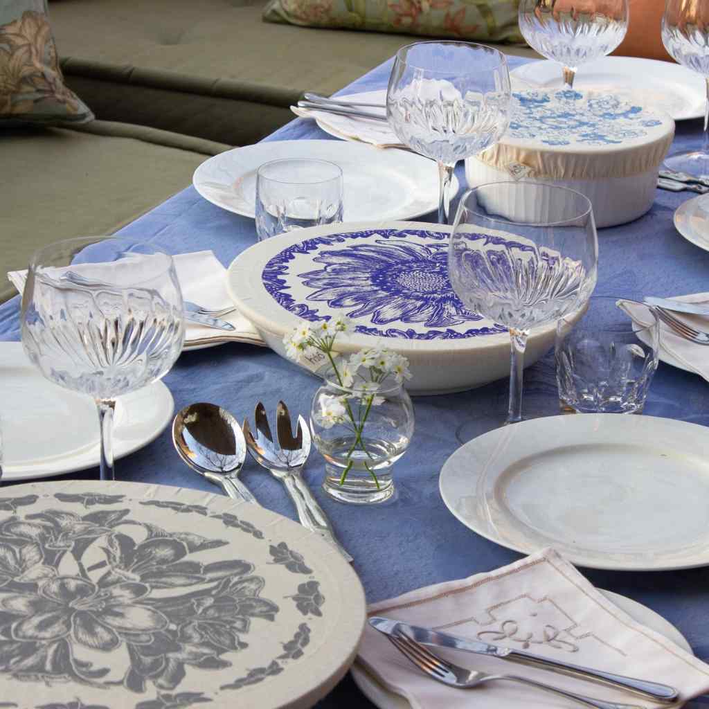 Elegant table setting with floral-patterned dishcovers and white plates, glasses, and cutlery on a blue tablecloth.