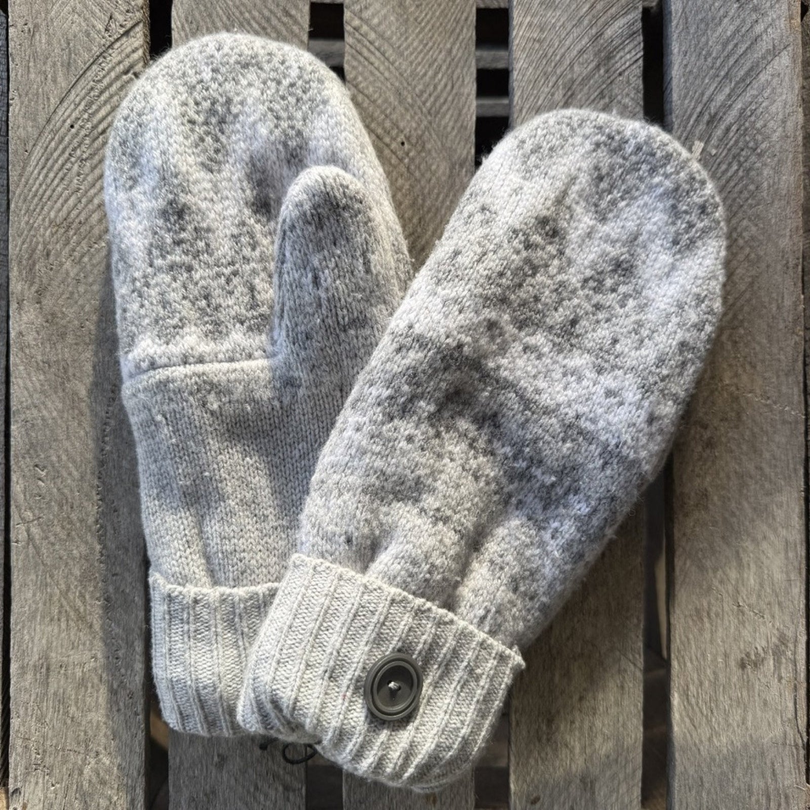 A pair of light gray knit mittens with a buttoned wrist closure, laid flat on a weathered wooden slatted surface.
