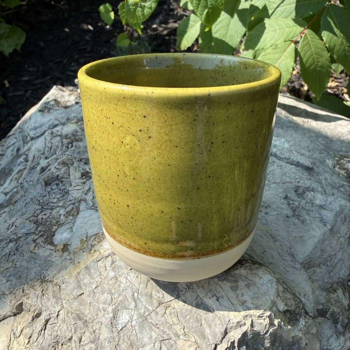 A handmade ceramic mug (or planter pot) with a speckled olive green glaze and a natural unglazed beige base, shown outdoors on a stone surface with green foliage in the background.