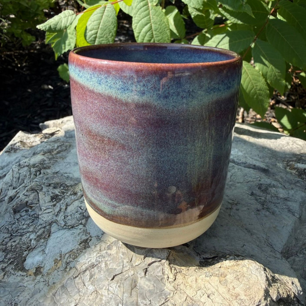 A handmade ceramic mug (or planter pot) with a glazed finish, featuring a gradient of purple, blue, and brown on the upper body, and a natural beige unglazed base, sitting on a stone surface with green foliage in the background.