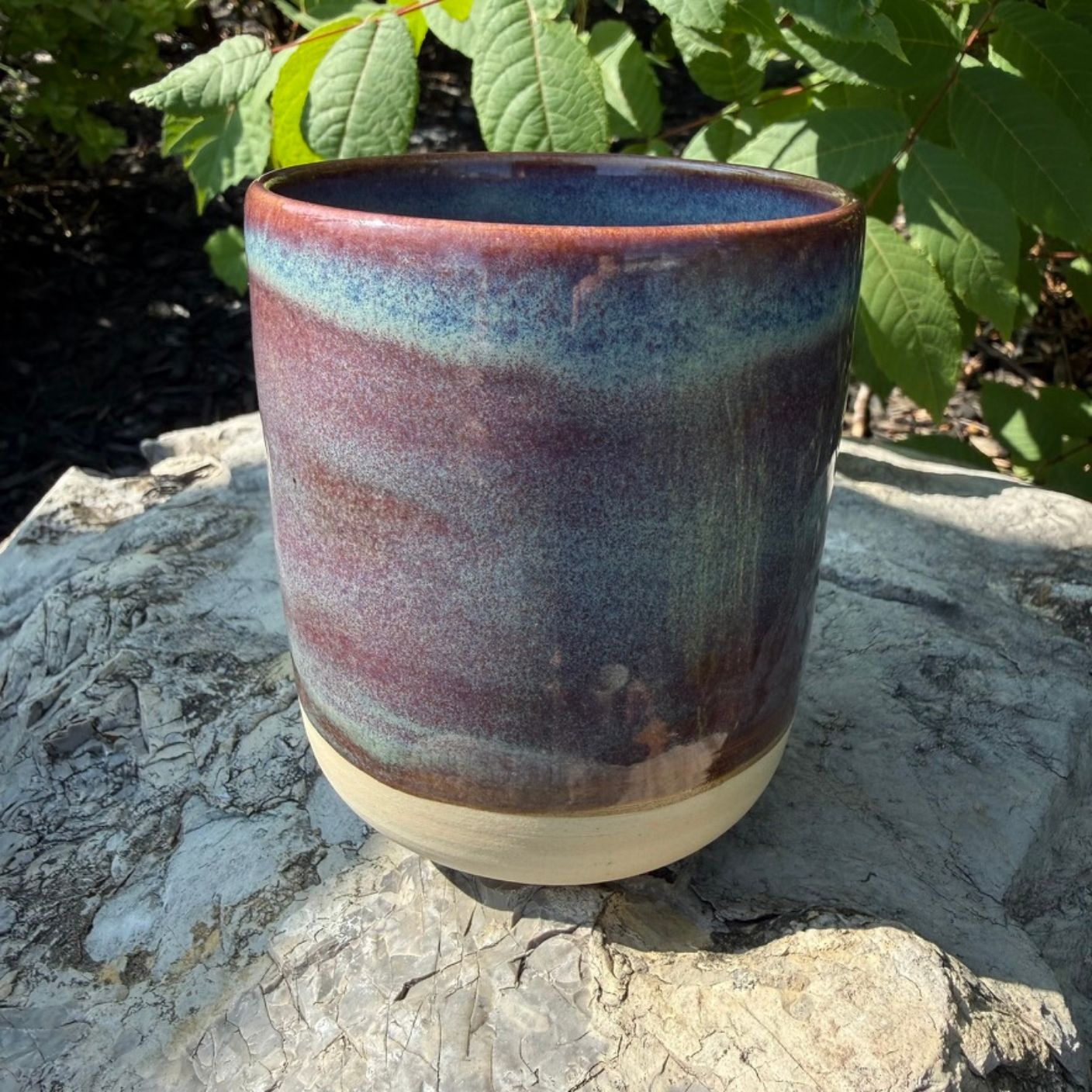A handmade ceramic mug (or planter pot) with a glazed finish, featuring a gradient of purple, blue, and brown on the upper body, and a natural beige unglazed base, sitting on a stone surface with green foliage in the background.