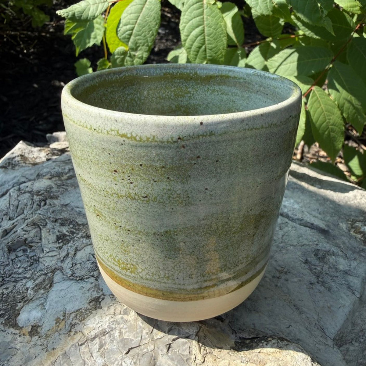 A handmade ceramic mug (or planter pot) with a rustic glaze in shades of sage green and beige, sitting outdoors on a stone surface with green foliage in the background.
