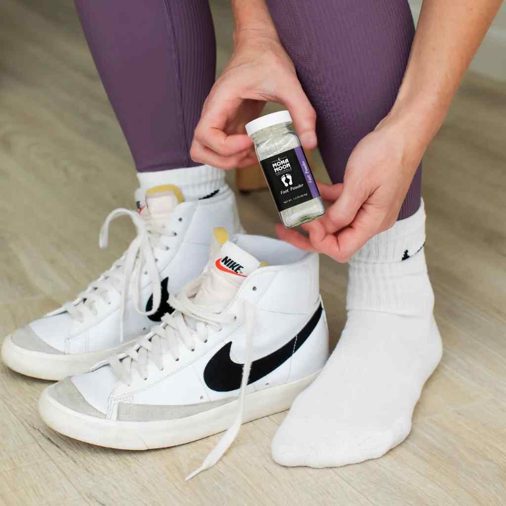 Person wearing white sneakers with a black Nike logo, holding a container of Mona moon talc-free foot powder, and standing on a wooden floor.
