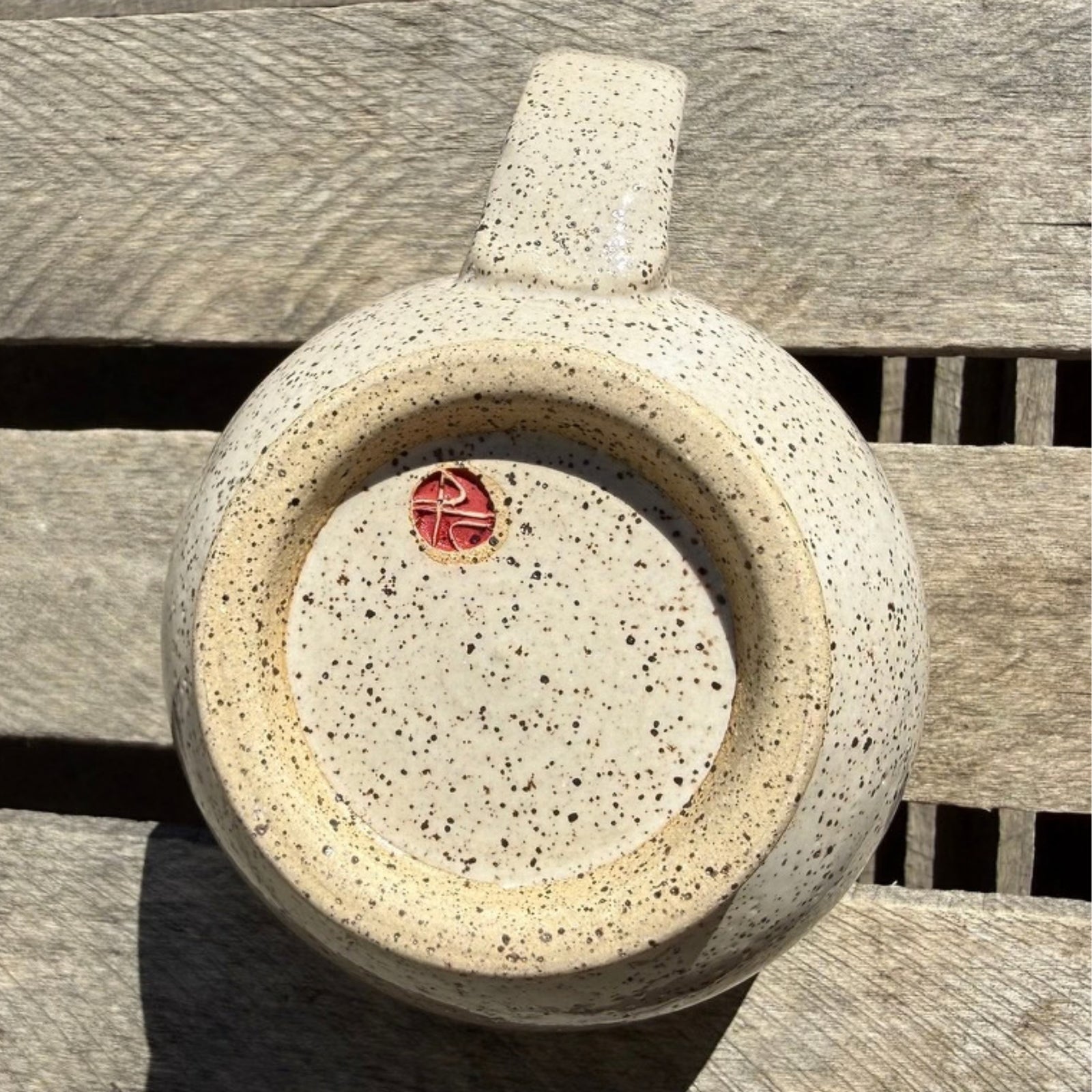 Bottom of a speckled beige handmade mug with maker's insignia showing PC.