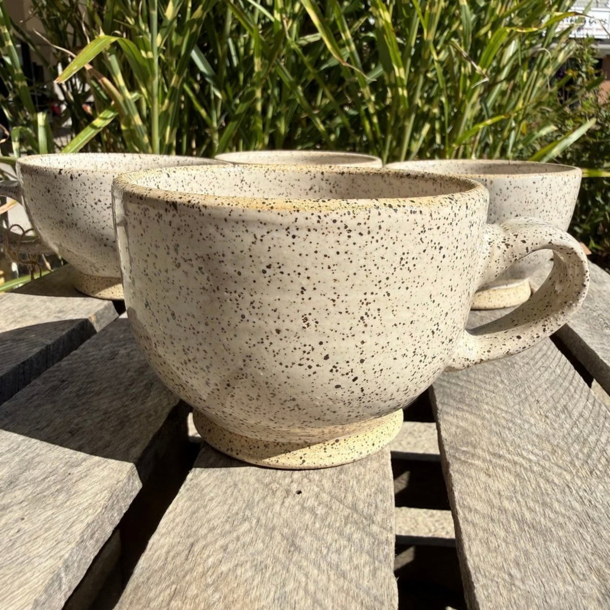 A speckled beige ceramic mug with a thick handle sits on a weathered wooden table outdoors, with other matching mugs and green foliage blurred in the background.