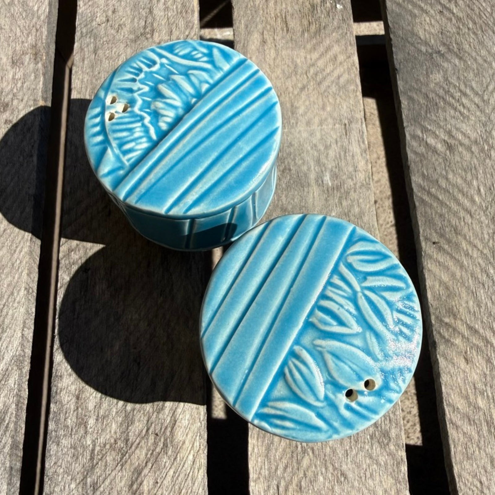 Two round turquoise blue ceramic salt and pepper shakers with textured leaf and stripe patterns, resting on a weathered wooden plank surface.