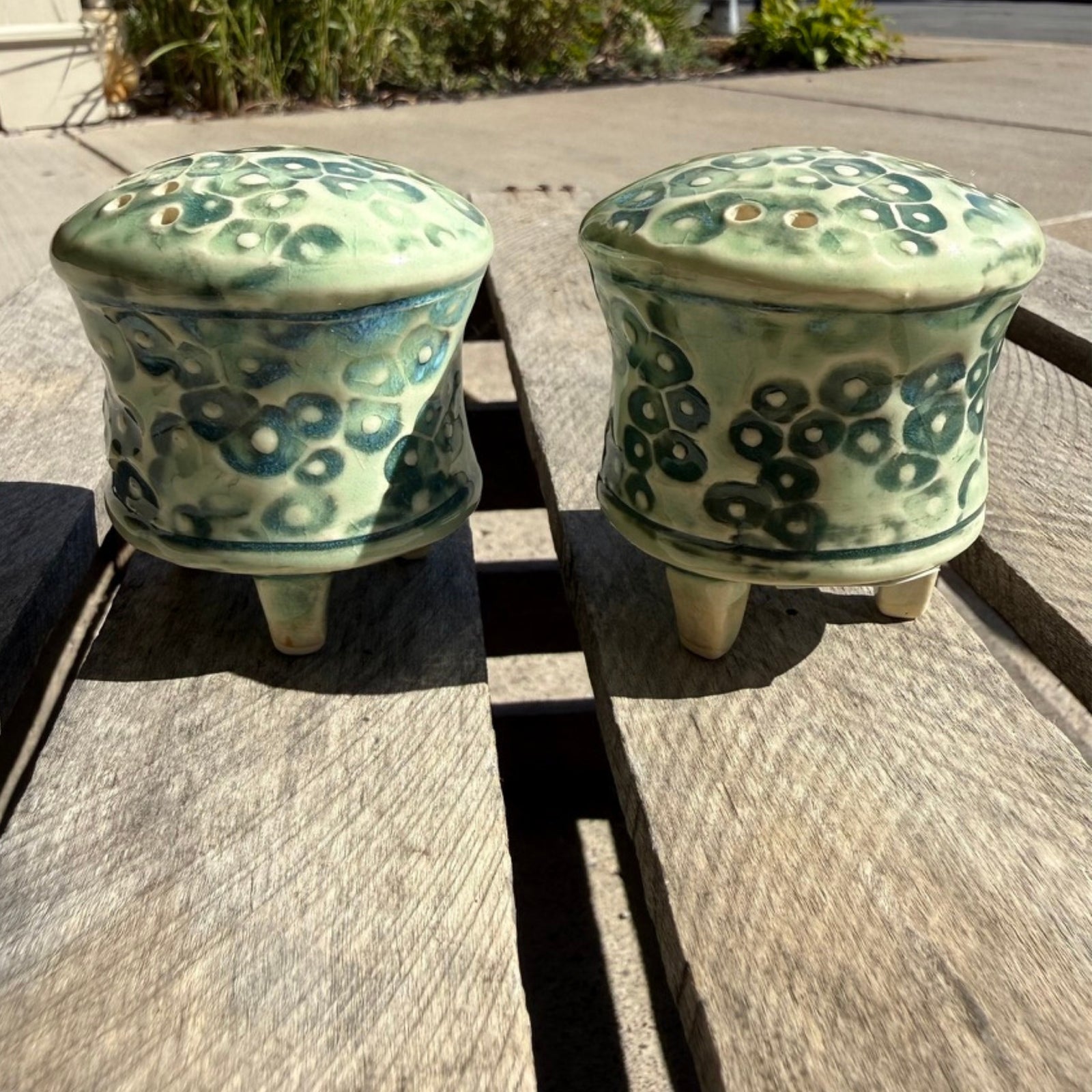 A pair of handmade ceramic salt and pepper shakers with a light green and dark green spotted pattern, sitting on a weathered wooden table outdoors.