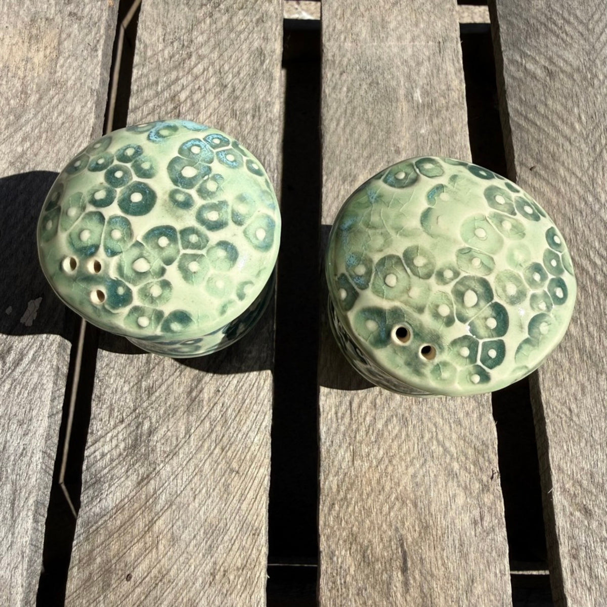 A pair of round ceramic salt and pepper shakers with a textured, bubble-like green glaze, resting on a weathered wooden plank surface.