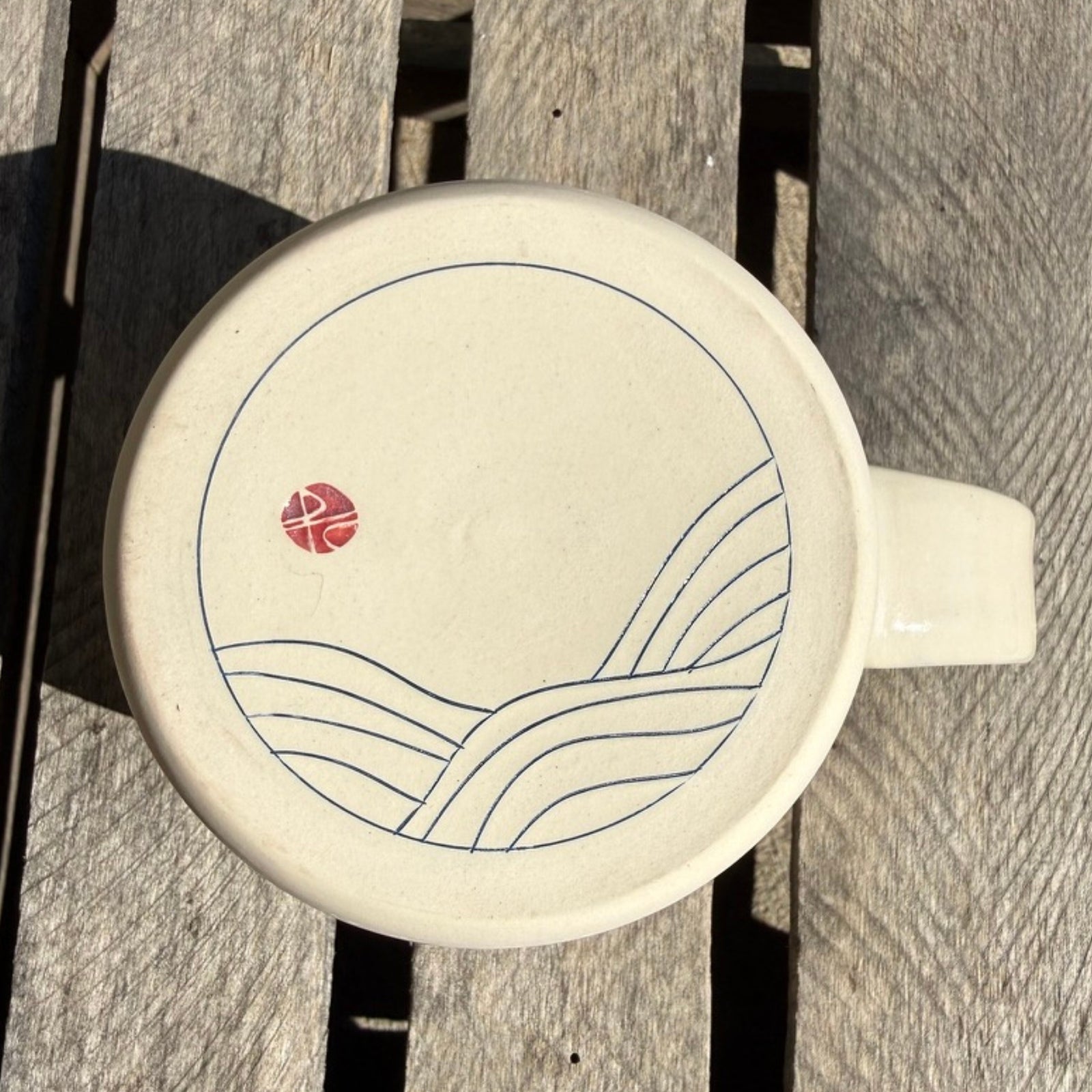 A round ceramic mug with a handle, shown from a top-down view of the bottom of the mug on weathered gray wooden slats. The mug is off-white with a minimalist blue line drawing of waves and a small red circular emblem.