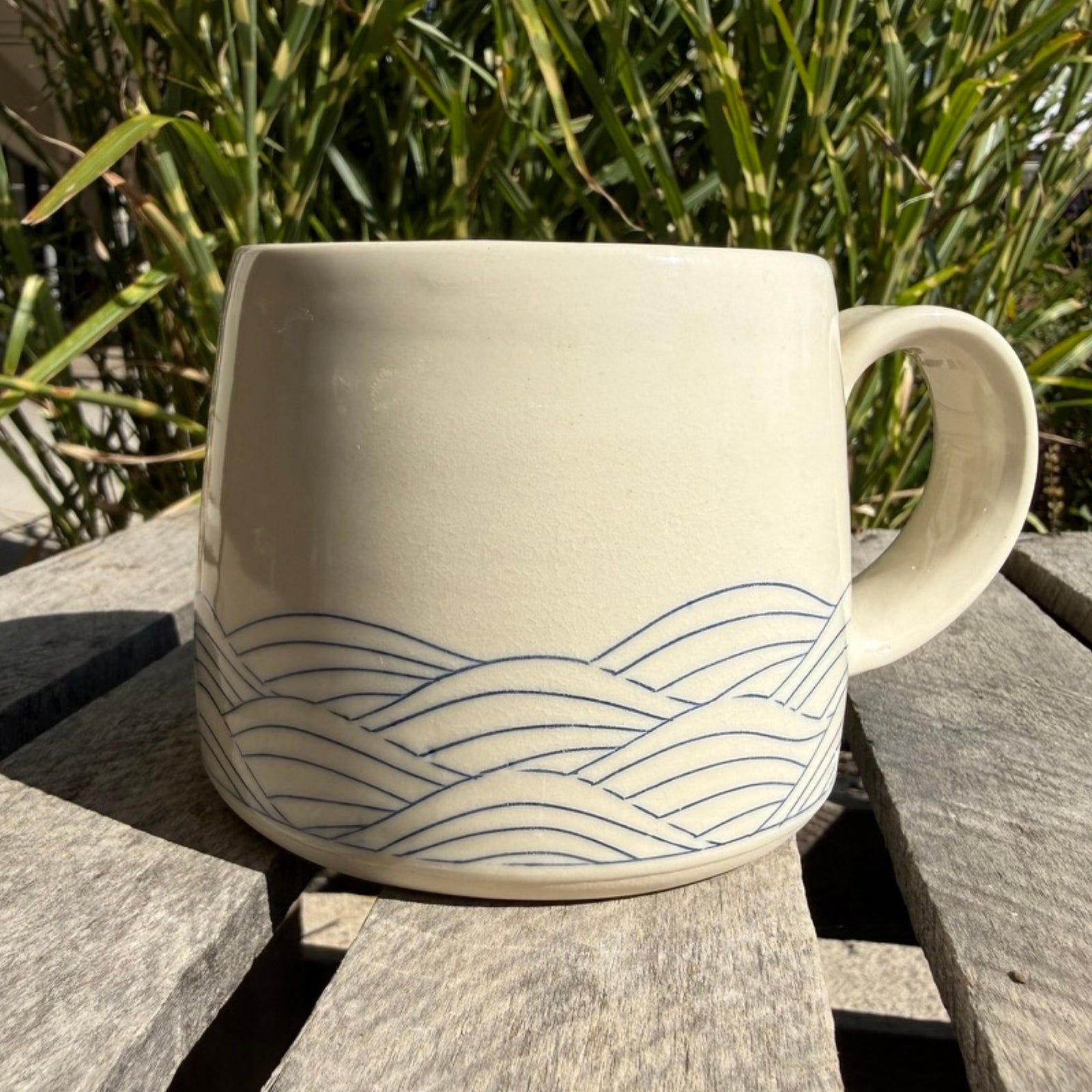 cream colored mug with blue wave glaze on wood crate with greenery in background.