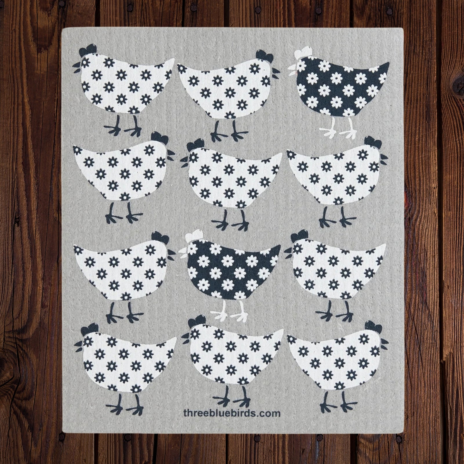 A swedish dishcloth featuring a repeating pattern of stylized chickens in black and white floral motifs on a light gray background, displayed flat against a dark wood plank backdrop. The text 'threebluebirds.com'
