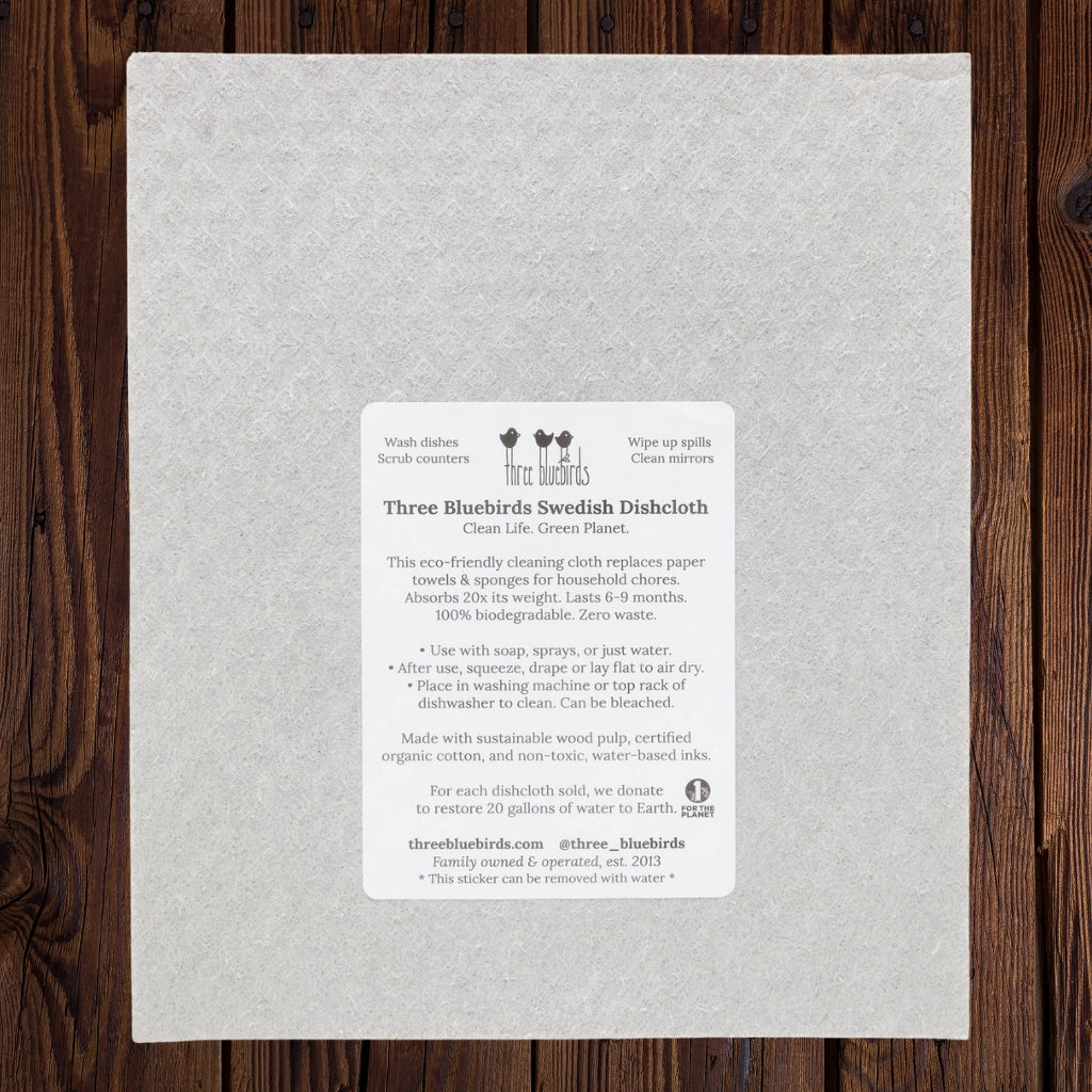 A square  light gray dishcloth with a subtle embossed pattern, featuring a white product information label on the front. The label reads 'Three Bluebirds Swedish Dishcloth' and includes product details and a small logo.