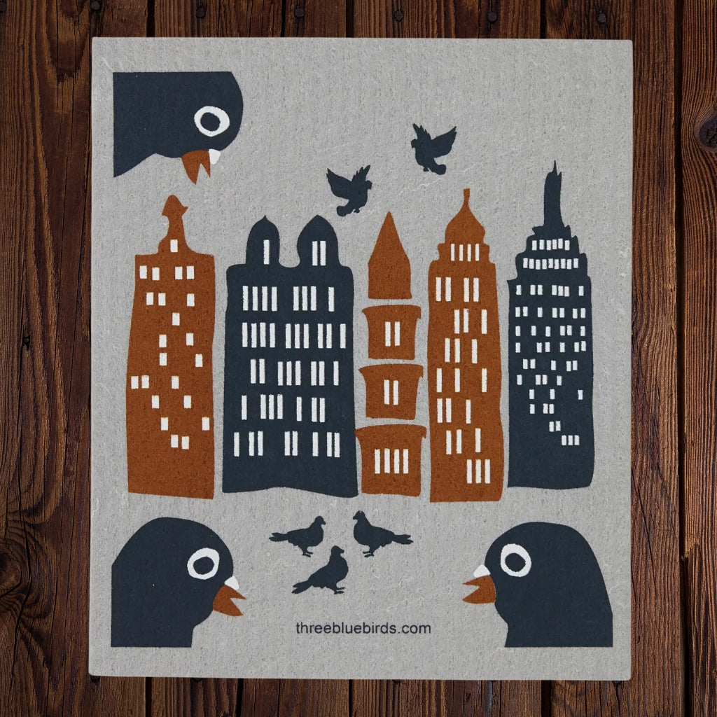 A square swedish dishcloth featuring stylized city buildings and birds on a light gray background, photographed flat against a dark wood surface. The artwork includes dark blue-gray birds with orange beaks, several dark blue-gray skyscrapers, and rust-brown skyscrapers with white window details.