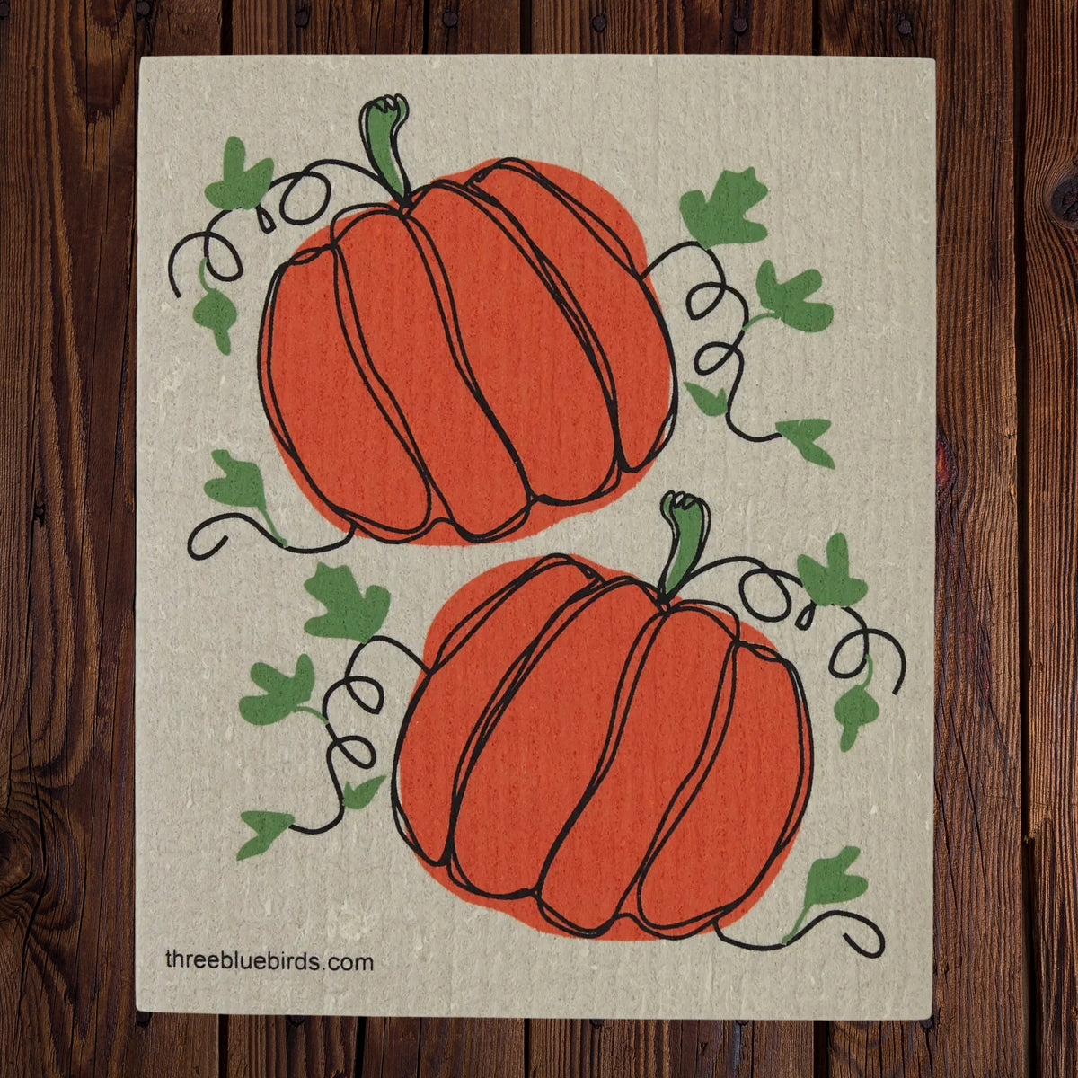 A square swedish dishcloth featuring two orange pumpkins with green leaves and vines on a textured beige background, displayed flat on a dark wood surface. The website 'threbluebirds.com'