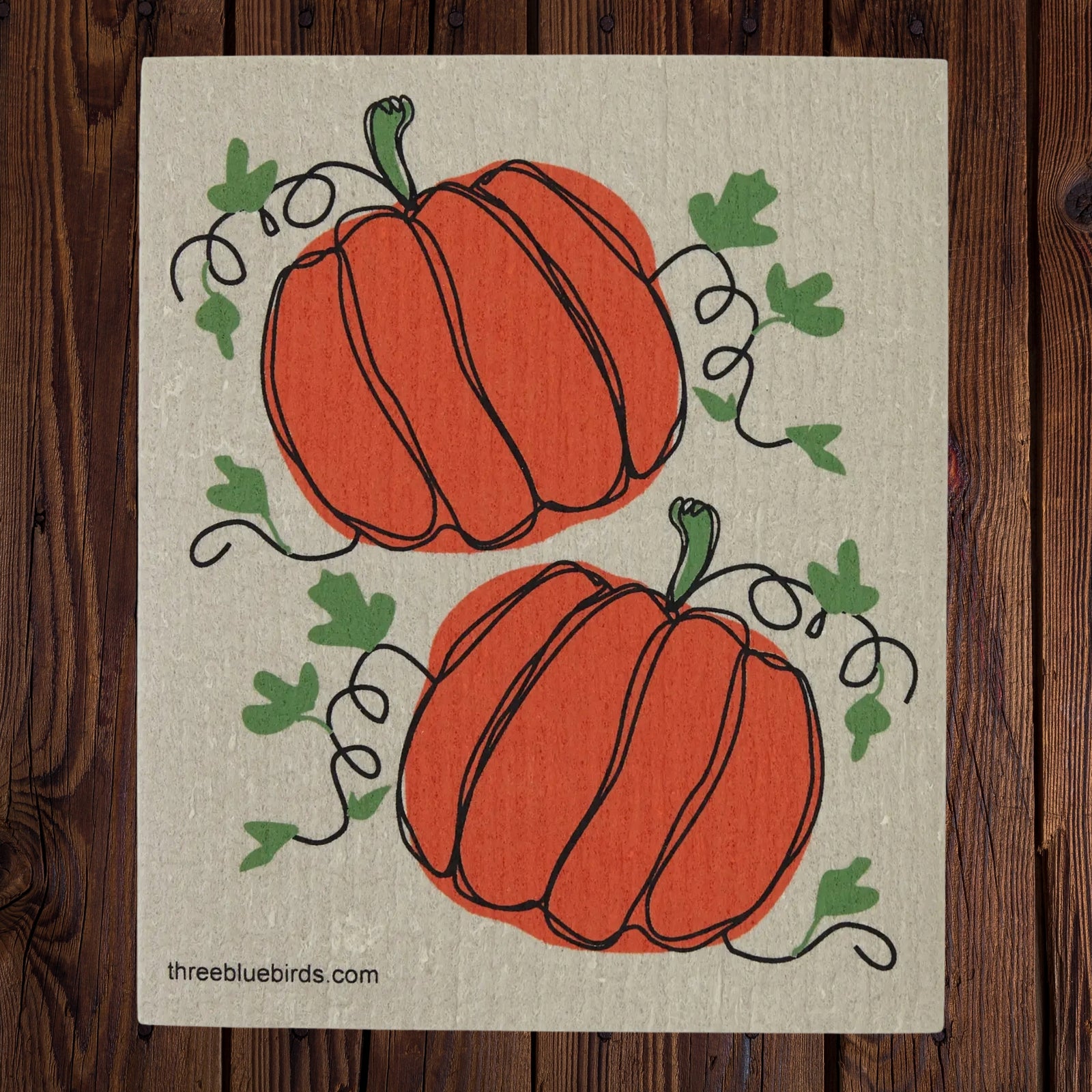 A square swedish dishcloth featuring two orange pumpkins with green leaves and vines on a textured beige background, displayed flat on a dark wood surface. The website 'threbluebirds.com'